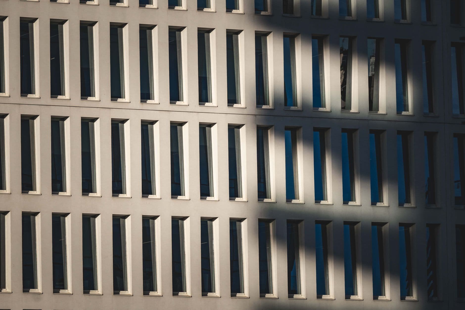 Regular grid of tall recessed windows on a light facade, partly crossed by a soft diagonal shadow.