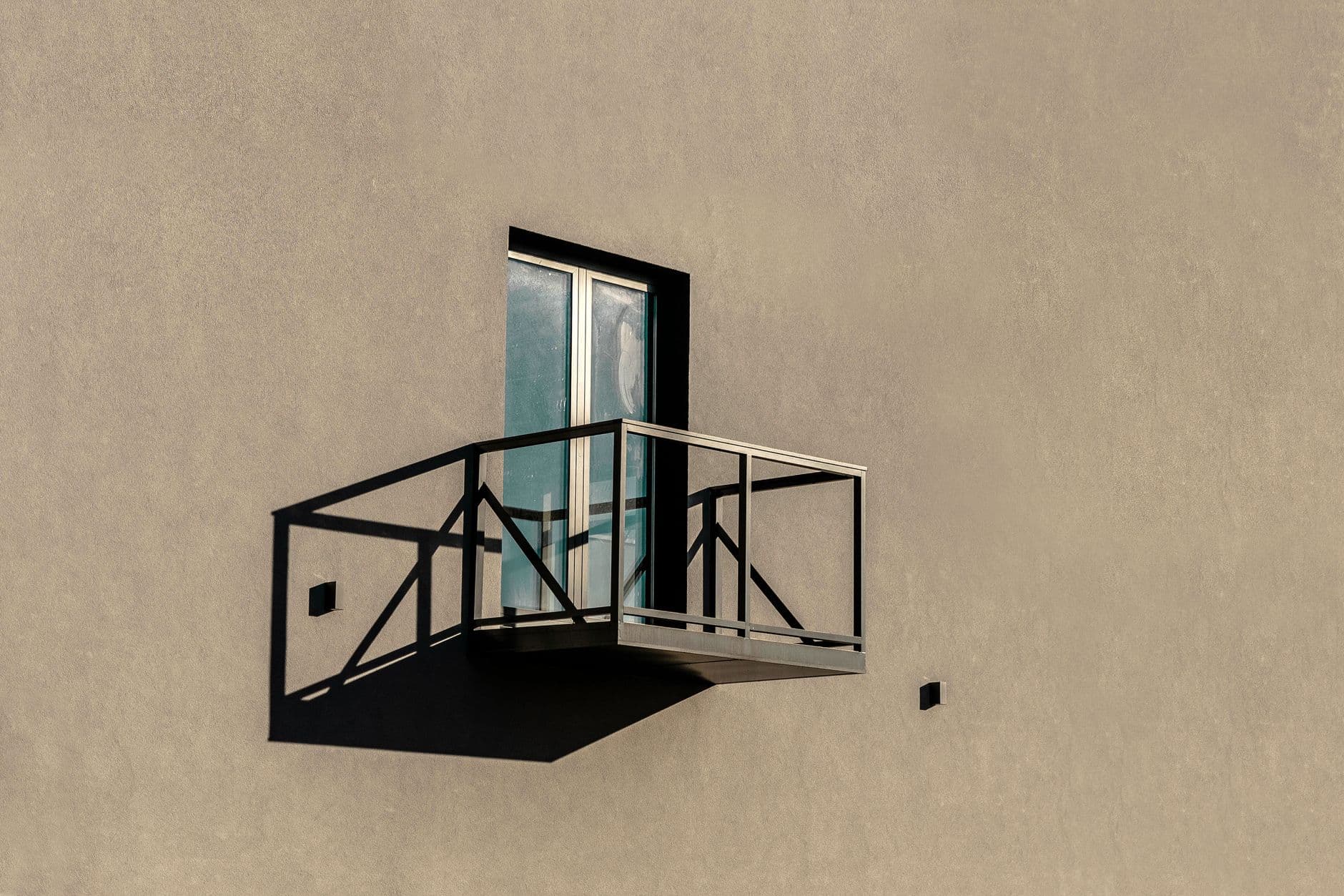 Single metal-railed balcony and window casting a sharp shadow on a plain beige rendered wall.