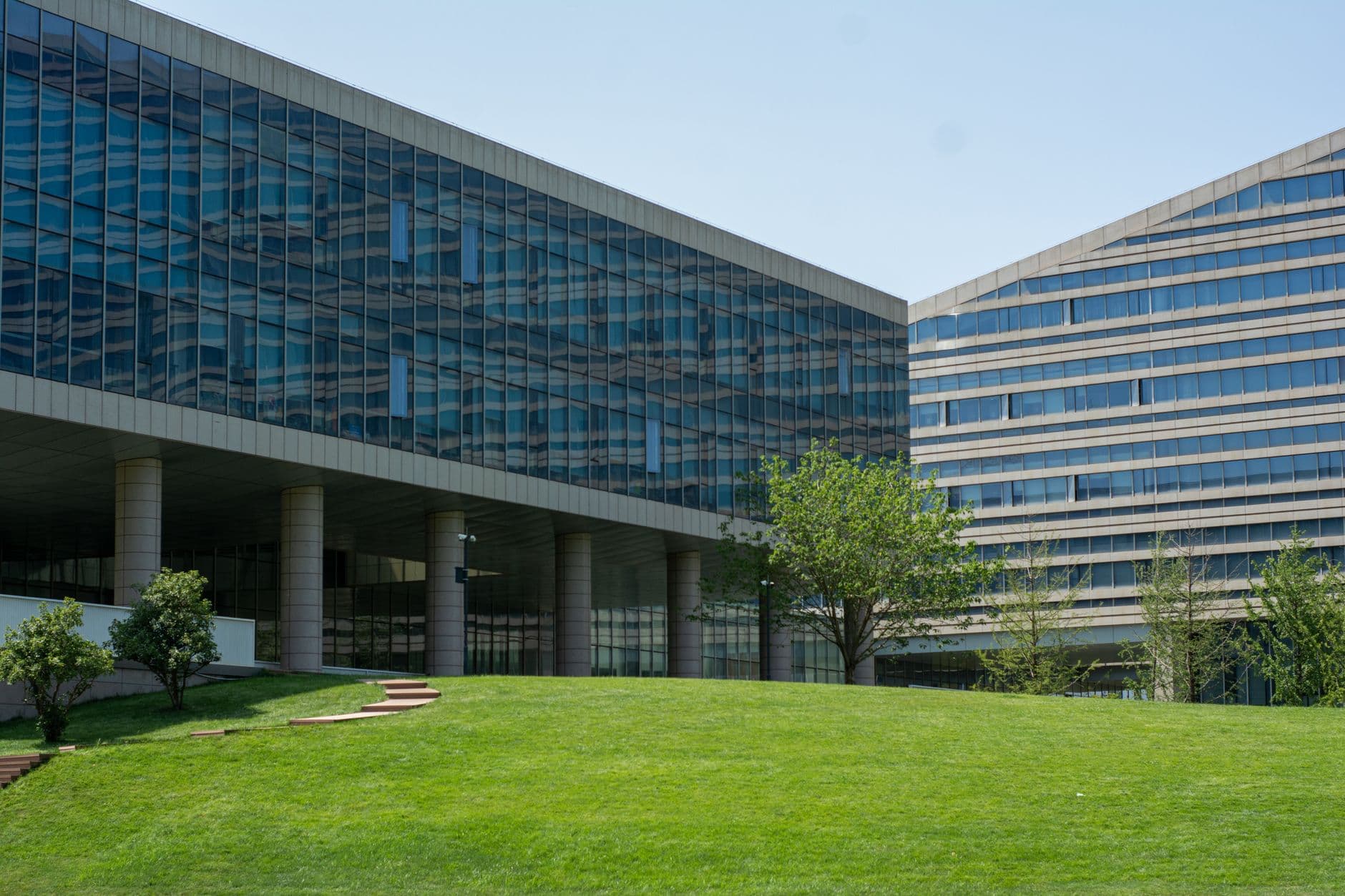 Two glass office buildings on a landscaped campus, with one block raised on columns above a lawn.